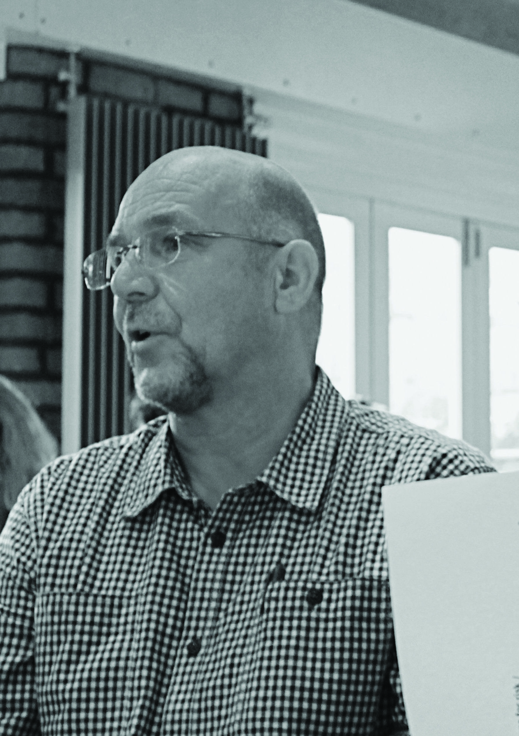 A man with glasses and a beard, wearing a checkered shirt, speaks during a meeting or workshop.