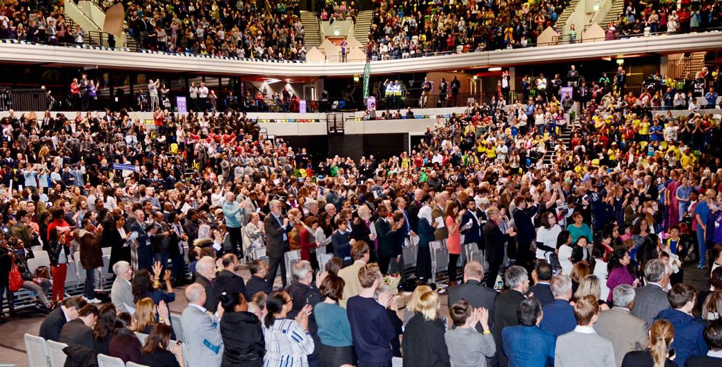 Large crowd of people standing and applauding in an indoor arena, indicating a substantial gathering or event.