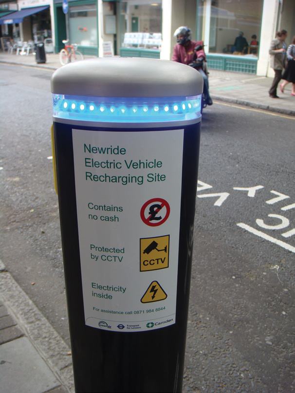 An electric vehicle recharging post on a street, displaying information about the Newride site, payment methods, security, and power details.