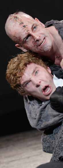 Two male actors with intense expressions, one bald with stitched makeup, grappling in a dramatic stage scene.