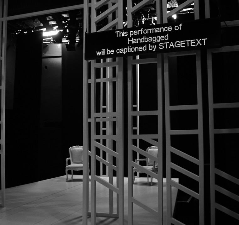 Black and white photo of a stage set with white lattice structures and two chairs. A sign reads: 'This performance of Handbagged will be captioned by STAGETEXT.'