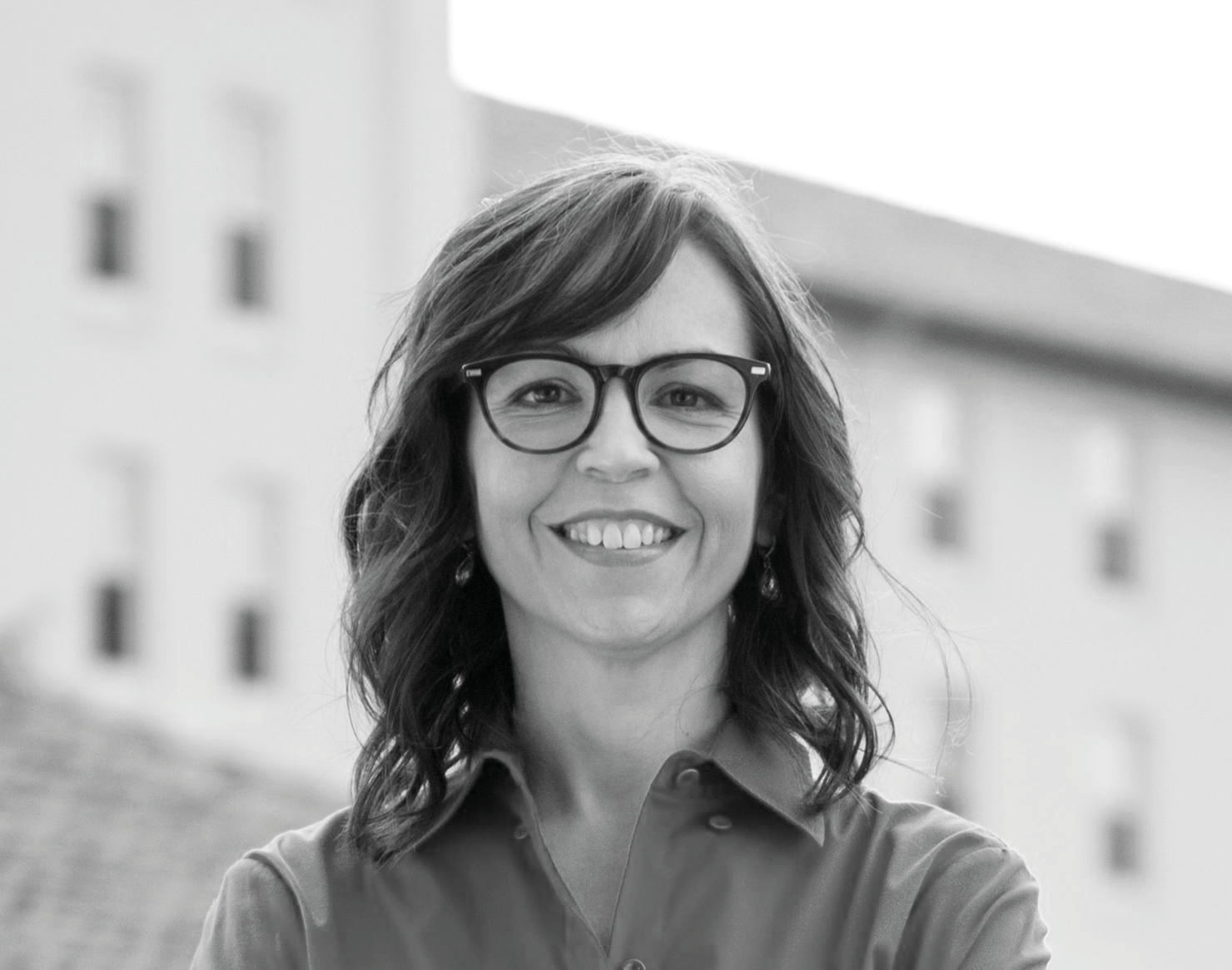 Black and white headshot of a smiling woman wearing glasses, with a blurred building in the background.