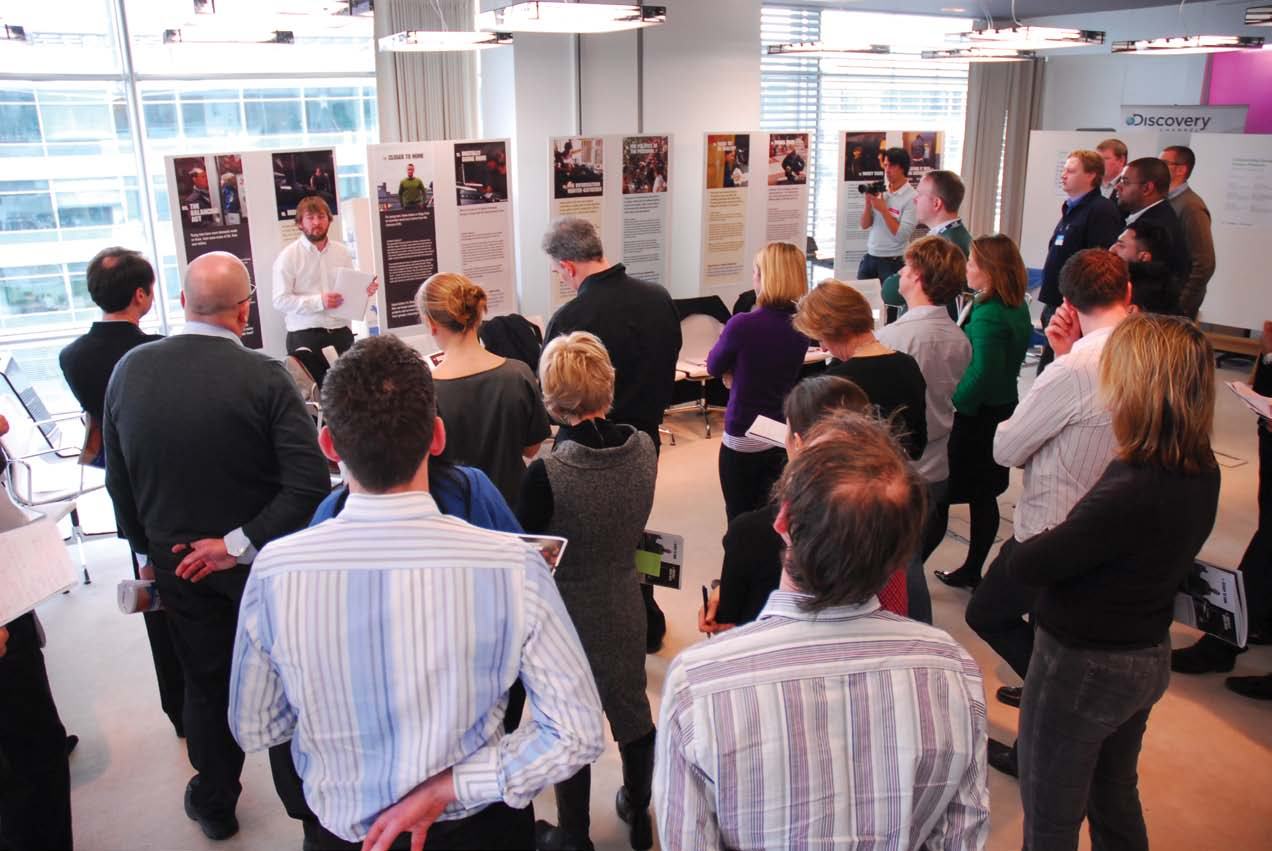 Group of people attending a presentation in a room with informational posters and a "Discovery" logo visible in the background.
