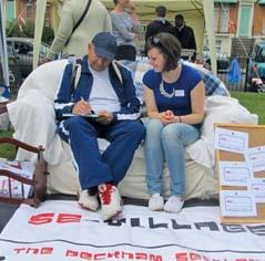 A man writing on a clipboard while sitting next to a woman on a couch outdoors, with a "SE-VILLAGE" banner in front, at a community event.
