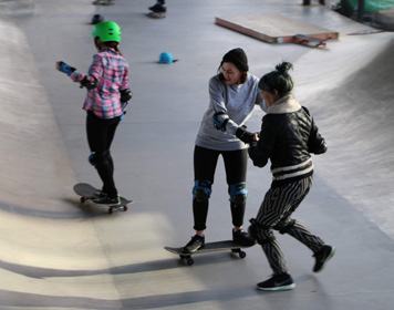 Two people skateboarding in a concrete skate park, one assisting the other, both wearing protective gear.