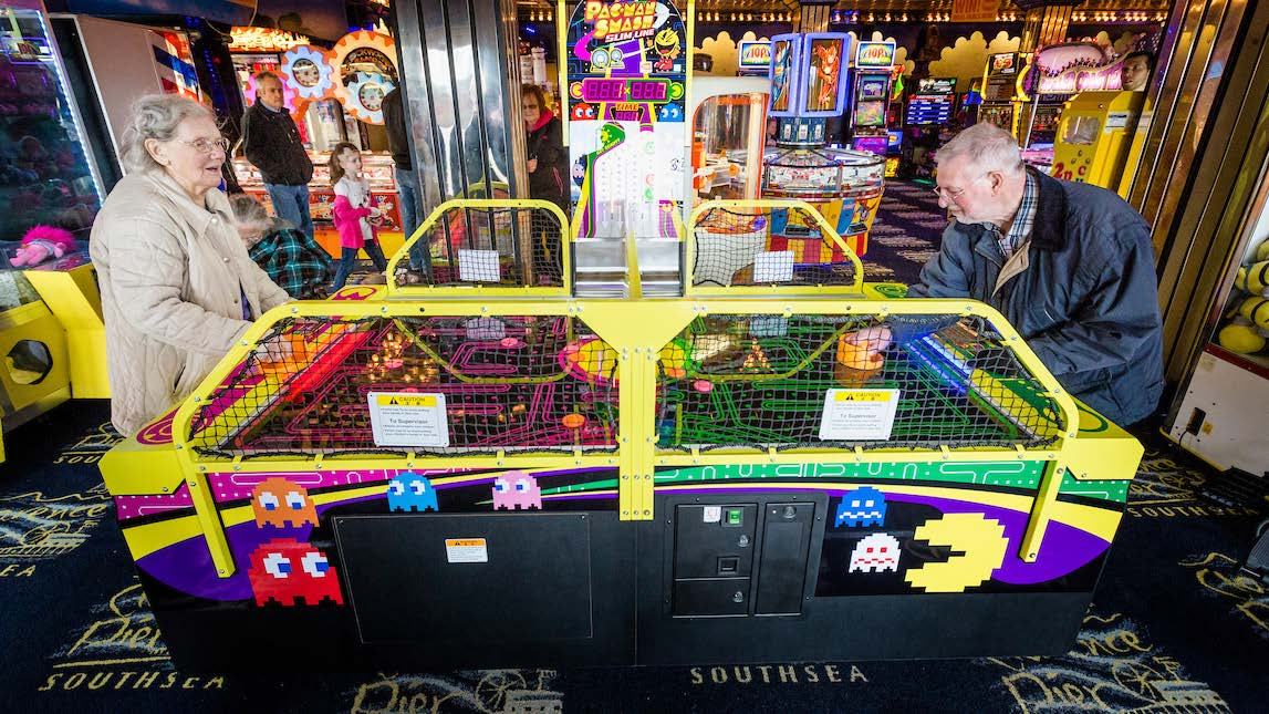 Two elderly people playing a Pac-Man themed arcade game in an amusement arcade.