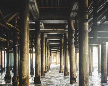 View from underneath a long wooden pier, showing numerous support pillars extending into the water, with waves on the ocean.