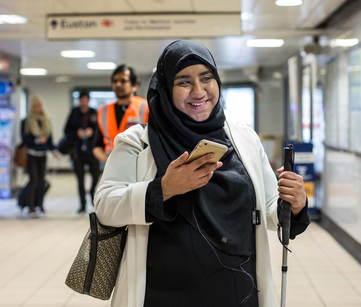 A smiling woman wearing a hijab, white jacket, and black dress, holding a smartphone and a white cane, walking in a brightly lit public transport station.