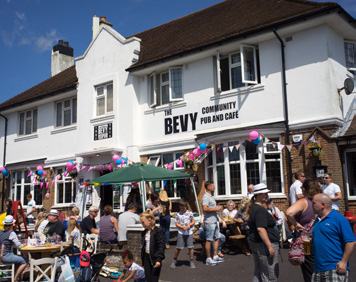 Exterior of "The Bevy Community Pub and Cafe" with many people gathered outside during a sunny event, decorated with balloons and bunting.