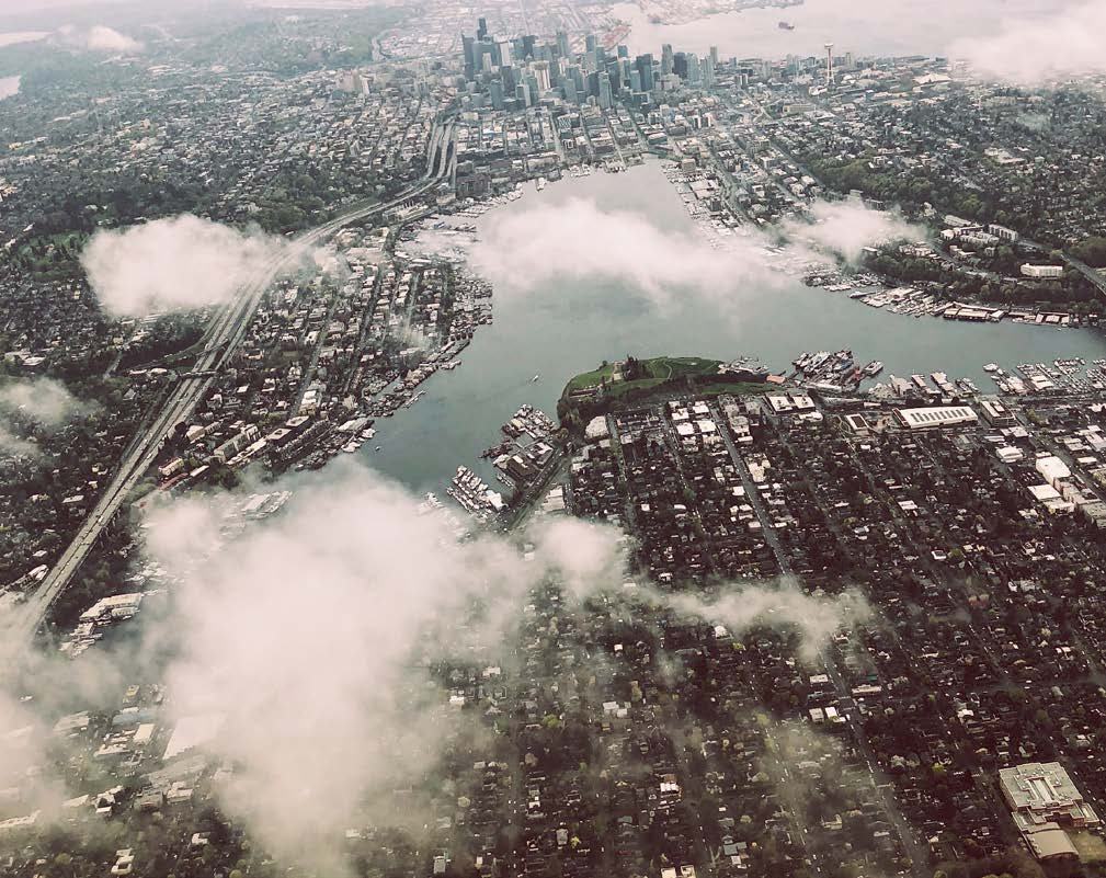 Aerial view of a sprawling city with a downtown skyline, a large body of water with boats, and multiple highways cutting through, partially obscured by clouds.