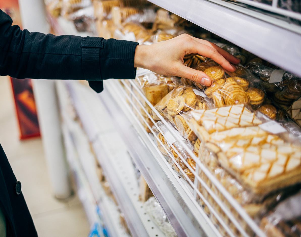 A person's hand reaches for packaged baked goods on a shelf in a supermarket aisle.