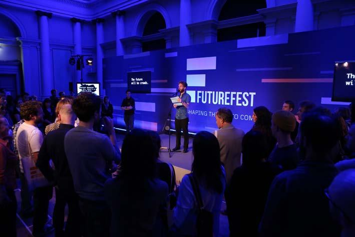 A speaker on stage addressing an audience at an event with a "FUTUREFEST" banner in a room lit with blue lights.