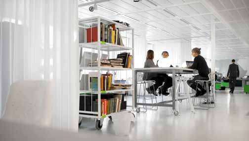 Wide shot of a modern, open-plan office space with people working at tables and a white bookshelf full of books.