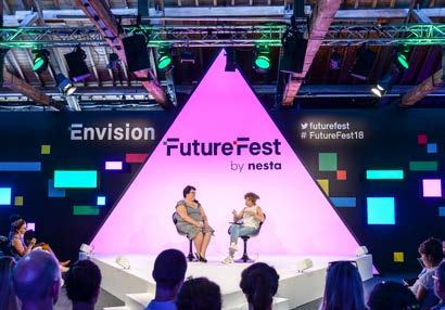 Two women on a stage with a large pink triangular backdrop featuring "Envision FutureFest by nesta" branding and an audience.