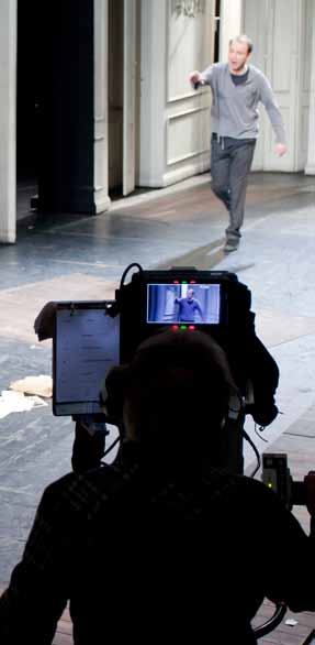Camera operator films an actor on stage in a modern interior set, with the actor visible on the camera's monitor.