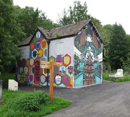 A building covered in colorful hexagonal murals and a large artistic figure, with a wooden signpost in front, likely in a park or cemetery.