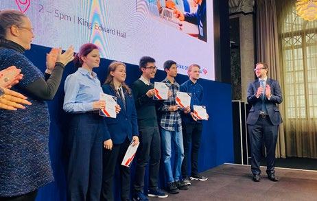 Group of students on stage receiving awards or certificates from an adult, with other adults applauding. A large screen is visible in the background.