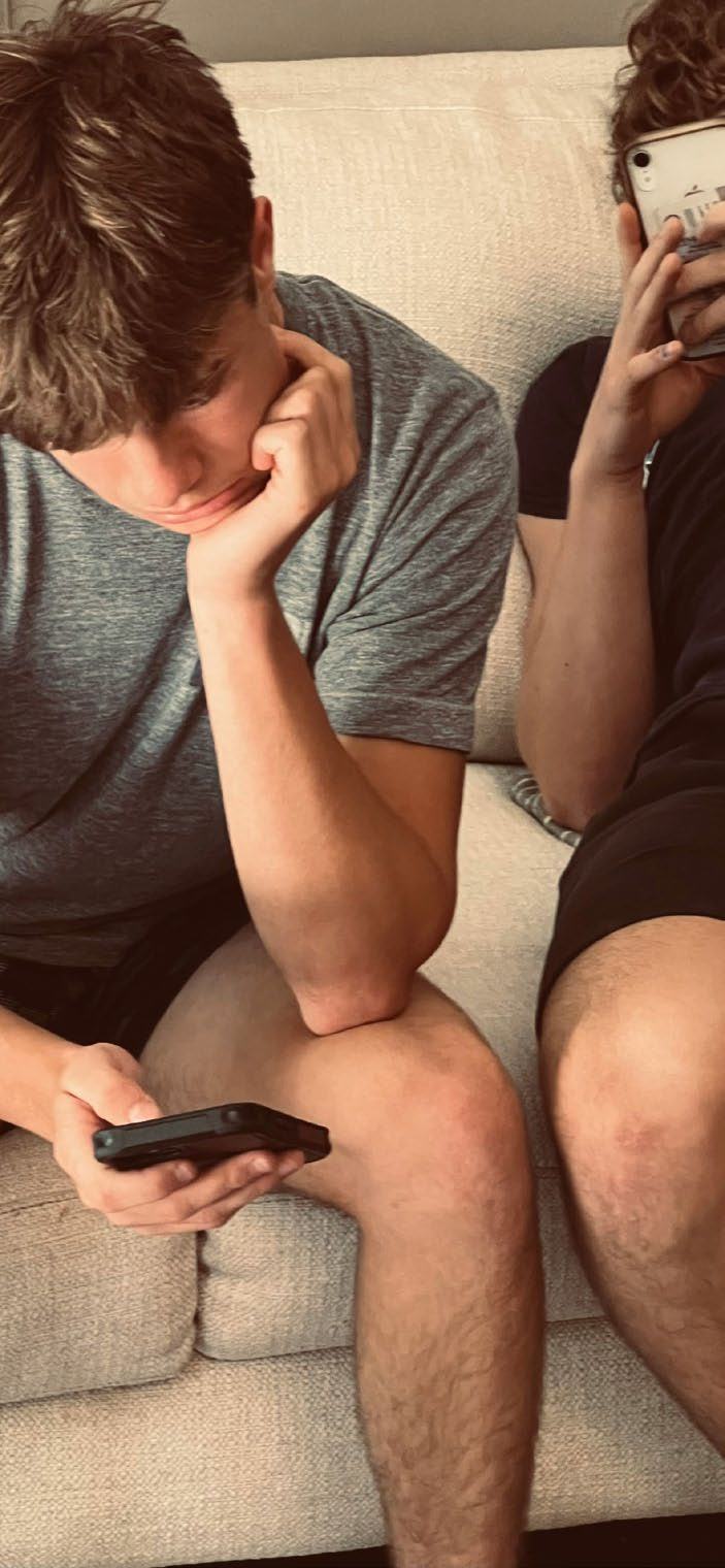 Two young men sitting on a couch, looking at their smartphones, with one resting his chin on his hand.
