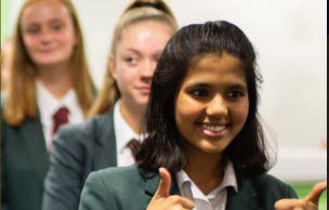 A smiling female student in a uniform gives two thumbs-up, with two other blurred students in the background.