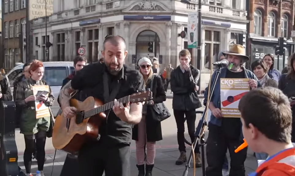 Street musicians performing, one playing guitar and another holding a sign requesting people to sign a petition related to CKO, with onlookers on a city street.