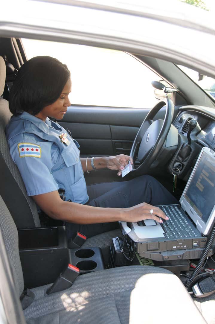A female police officer in uniform sits in a patrol car, typing on a mounted laptop and holding a card.