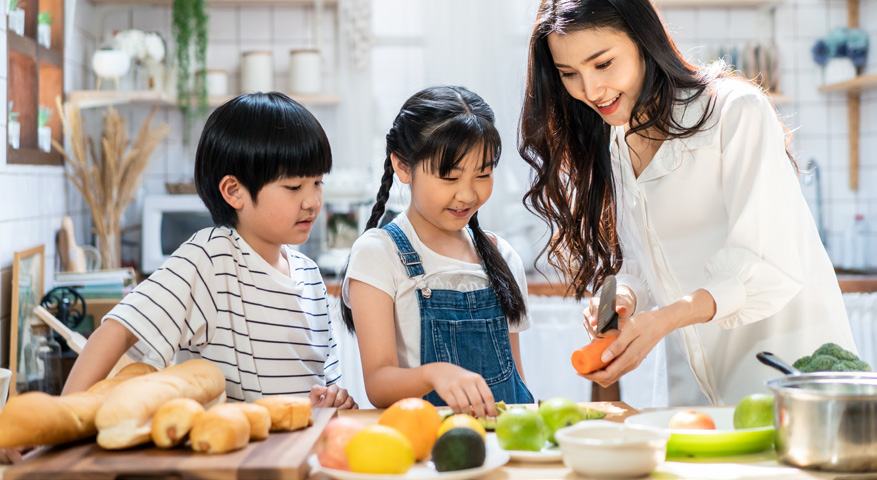 A mother and two children preparing food with fresh fruits and vegetables in a brightly lit kitchen.