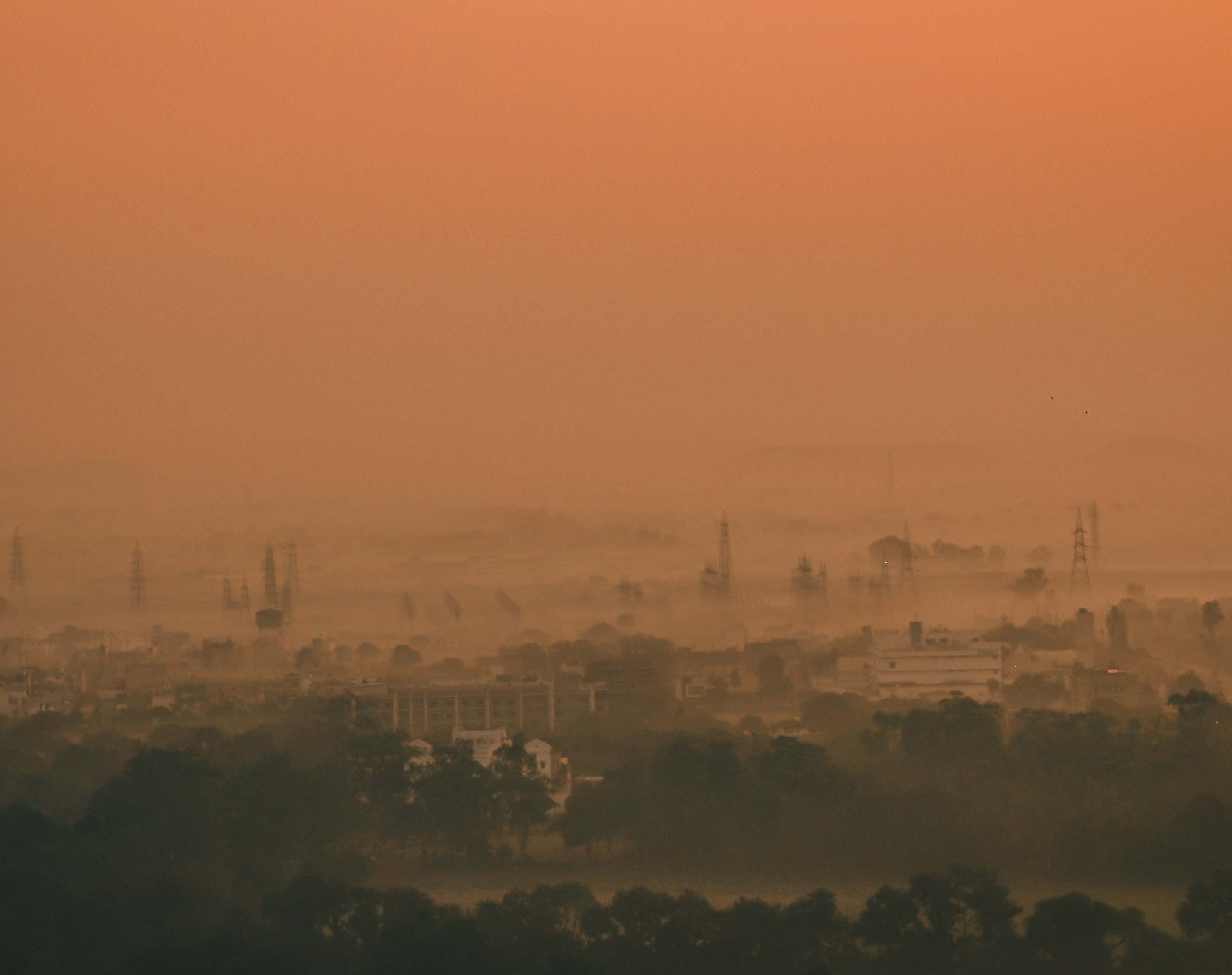 Hazy, reddish-orange landscape showing a city skyline with buildings and transmission towers obscured by fog or smog, indicating atmospheric conditions.