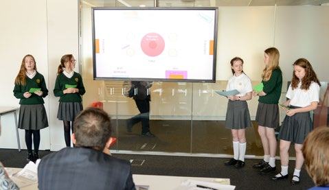 Students in uniform present in a room, with a large screen behind them showing a circular diagram.