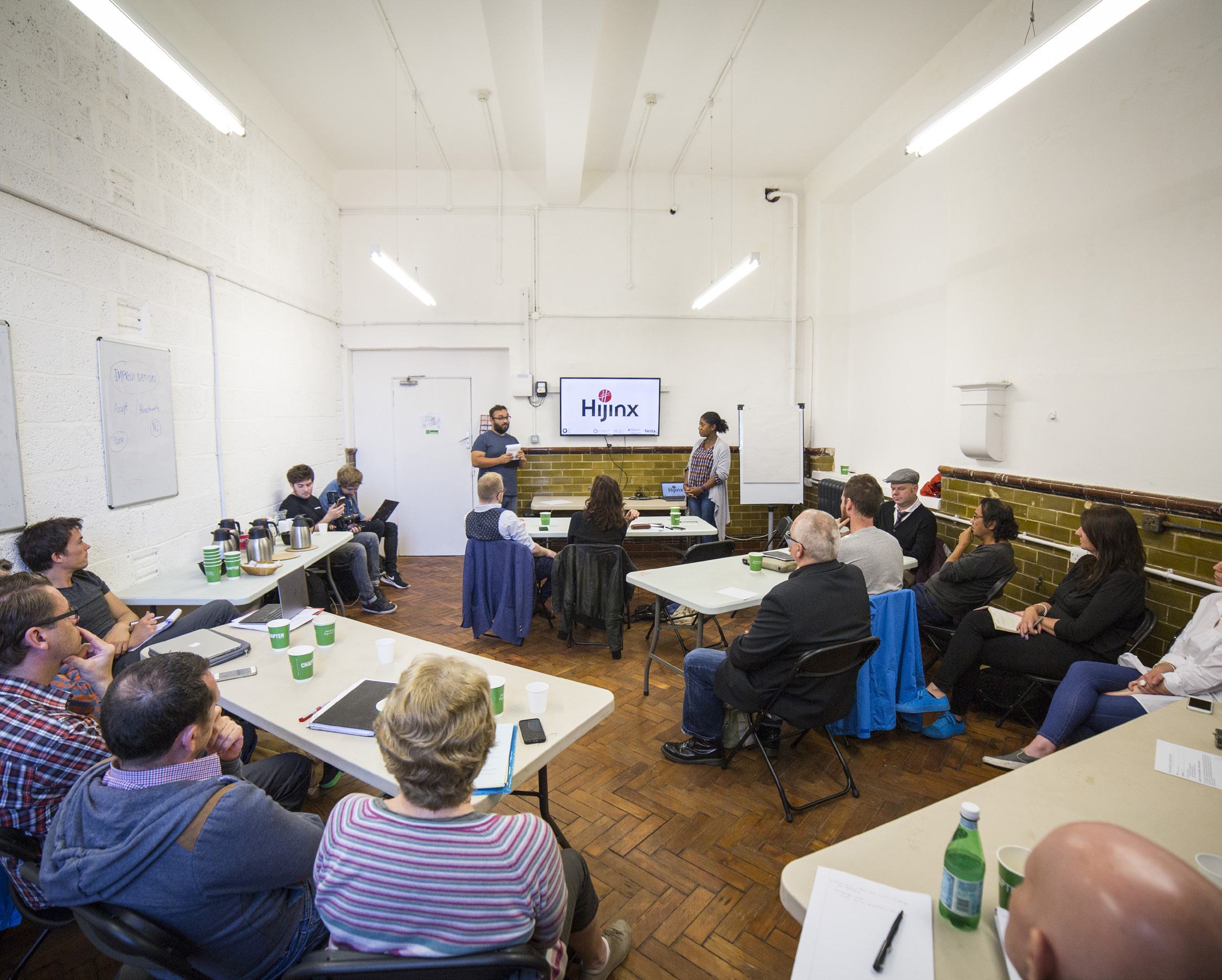 A wide shot of a workshop room with many participants seated at tables, facing a screen displaying the "Hijinx" logo.