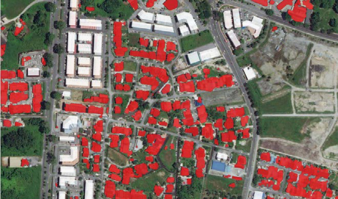 Aerial satellite view of an urban residential area with roads, buildings, and green spaces, where buildings are highlighted in red.