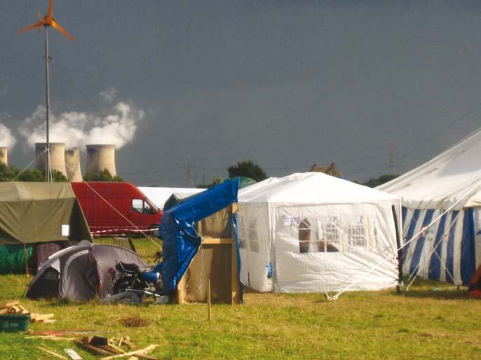 Tents and a wind turbine in a field with industrial cooling towers emitting steam in the background.