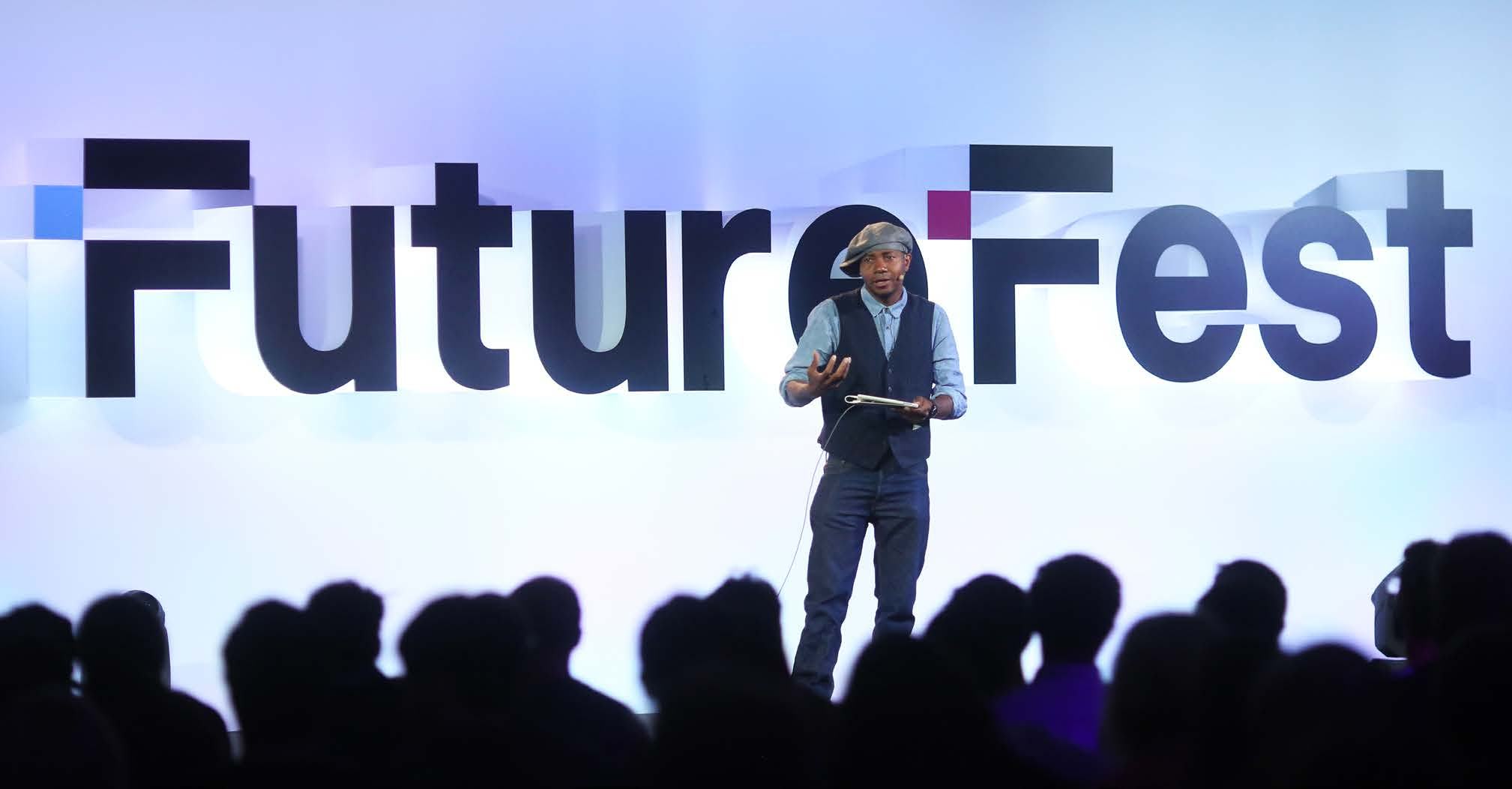 A man speaking on stage at a "FutureFest" event, gesturing with his hands, with an audience silhouetted in the foreground.