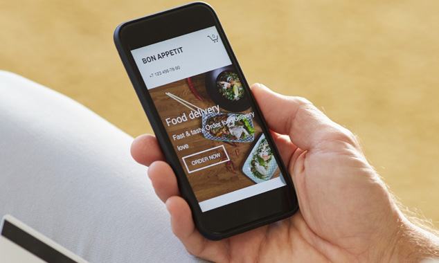 A person holding a smartphone displaying a food delivery app with "Bon Appetit" and "Food delivery" text and images of prepared dishes.