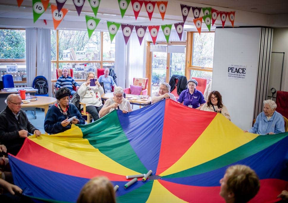 Elderly people and caregivers engaging in a group activity with a large colorful parachute indoors under "10TH ANNIVERSARY" bunting.