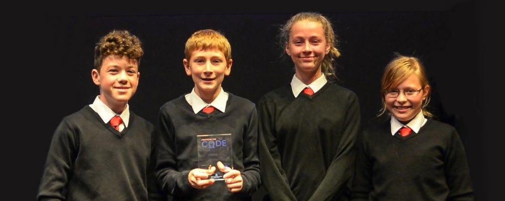 Four smiling students in uniform, two boys and two girls, stand on a stage. One boy holds a clear award trophy.