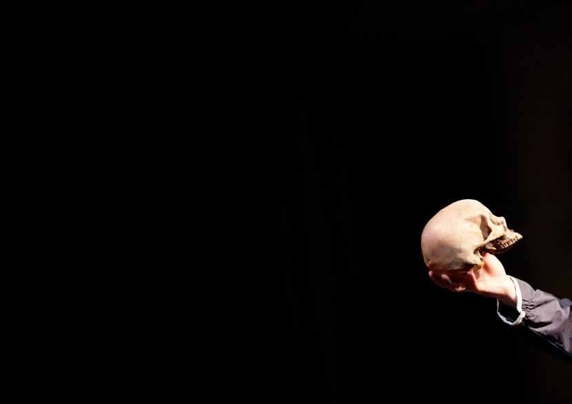 A hand holds a human skull against a stark black background, a classic theatrical prop.