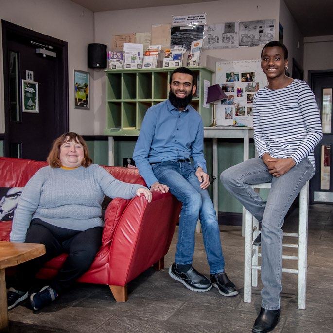 Three individuals, two men and one woman, pose smiling in a casual room with a red sofa and shelves in the background.