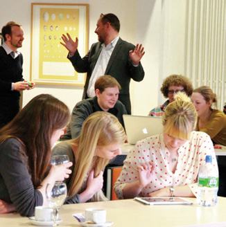 A group of people collaborating around a table in a meeting or classroom setting, with one man standing and gesturing.