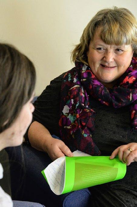 A woman with a colorful scarf smiles while talking to a person, holding a folded green document.