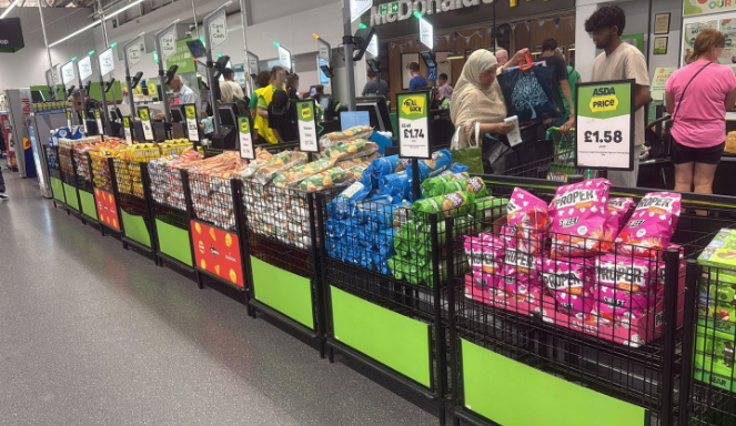 Supermarket checkout aisle showing a large display of crisps and snacks with "ASDA price" tags, and customers in the background.