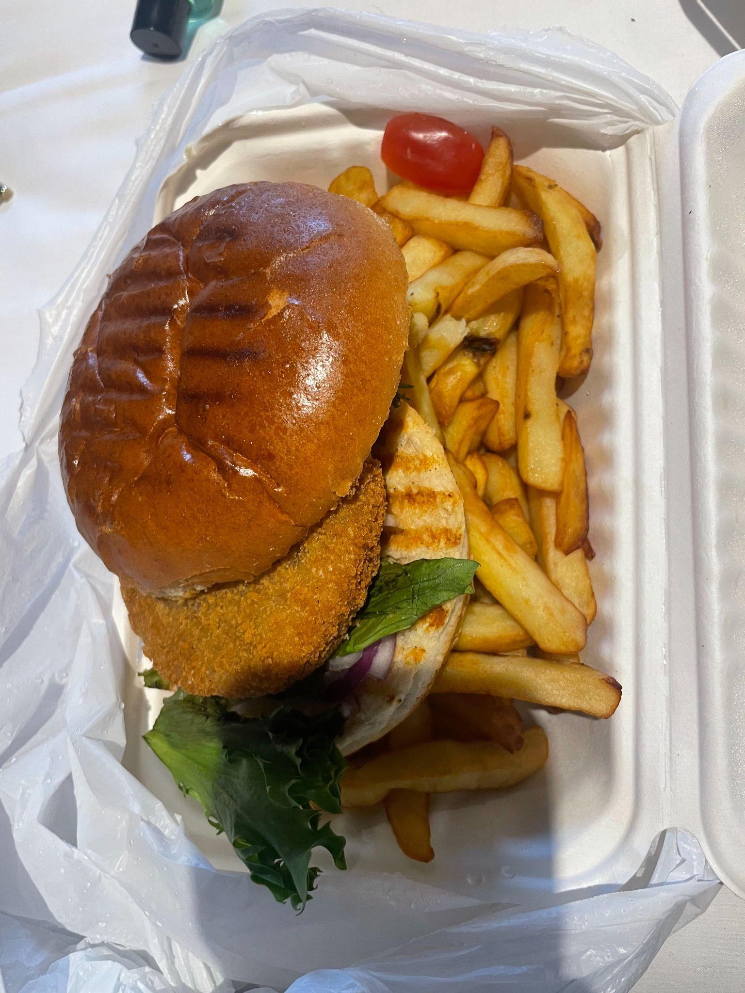 Takeaway box with a grilled chicken burger, fries, and a small cherry tomato.