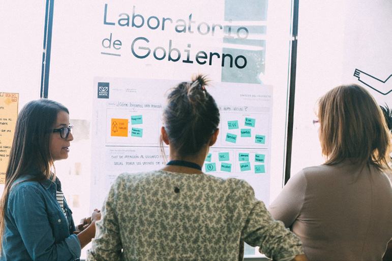 Three women discussing content on a large whiteboard covered with colorful sticky notes and diagrams, indicating a collaborative problem-solving session.