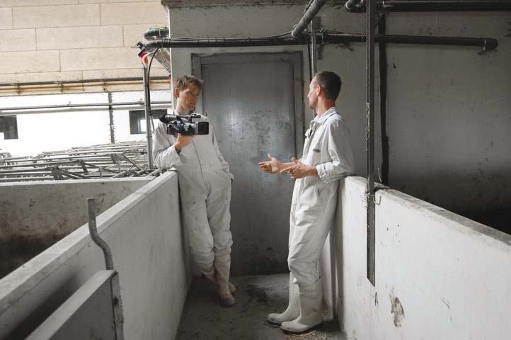 Two men in white coveralls, one filming with a video camera, talking in an indoor industrial setting.