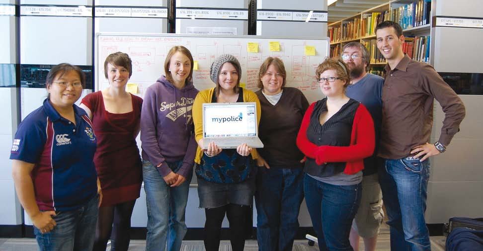 Diverse team of seven people standing in front of a whiteboard, one holding a laptop displaying 'mypolice'.