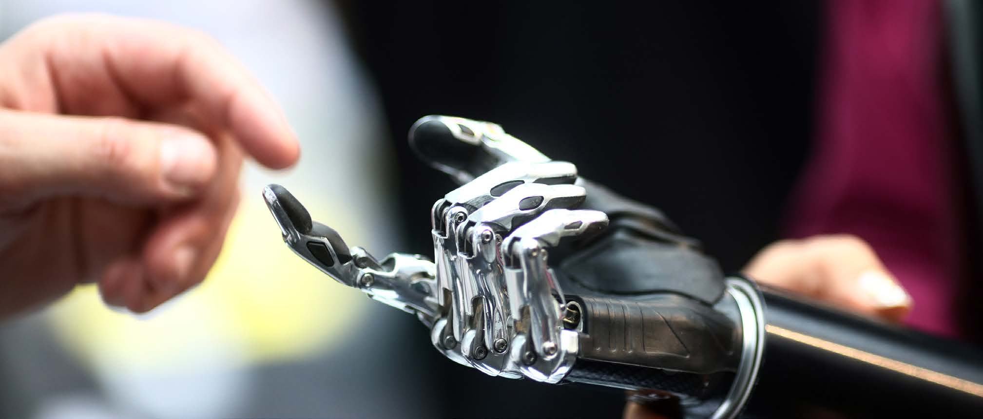 A human hand lightly touches the intricate fingers of a black and silver robotic prosthetic hand, suggesting interaction or technology.