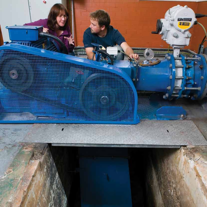 A man and a woman inspecting a large blue industrial motor and pump system in a utility room with an open pit.