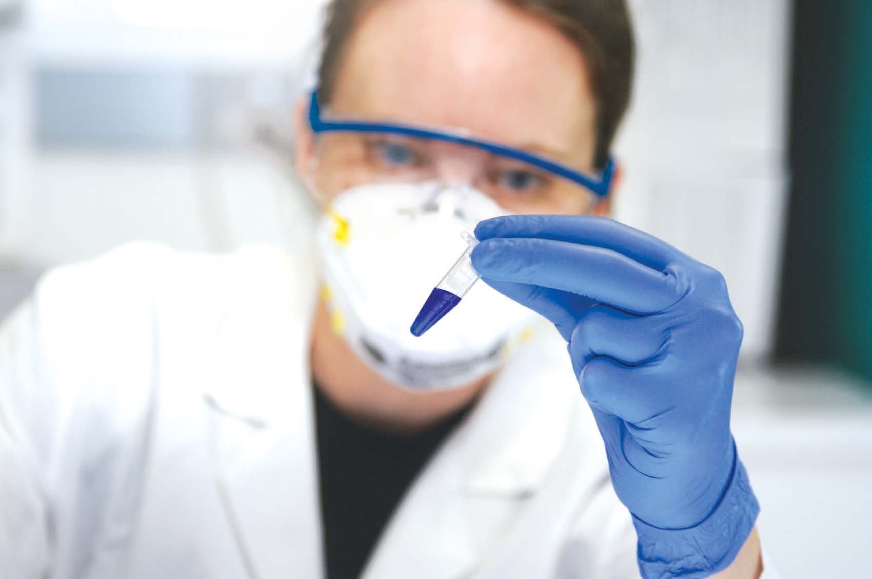 A scientist in safety goggles and mask holds a small vial with blue liquid, symbolizing research or laboratory work.