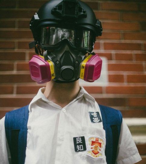 Person in school uniform wearing a tactical helmet, goggles, and a gas mask with pink filters, against a brick wall background.