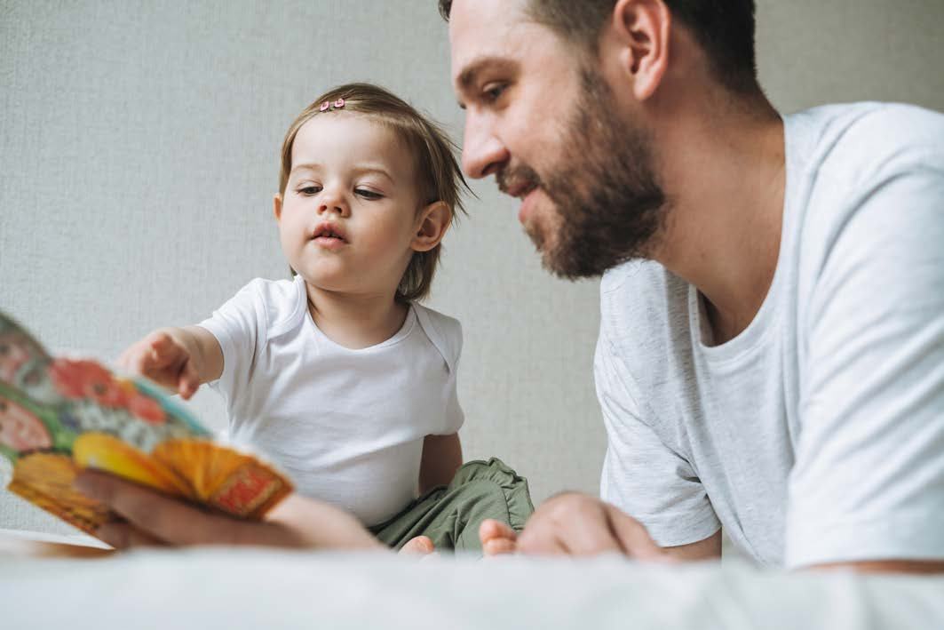 A father and young child in white shirts looking at a colorful picture book together, sharing a reading moment.