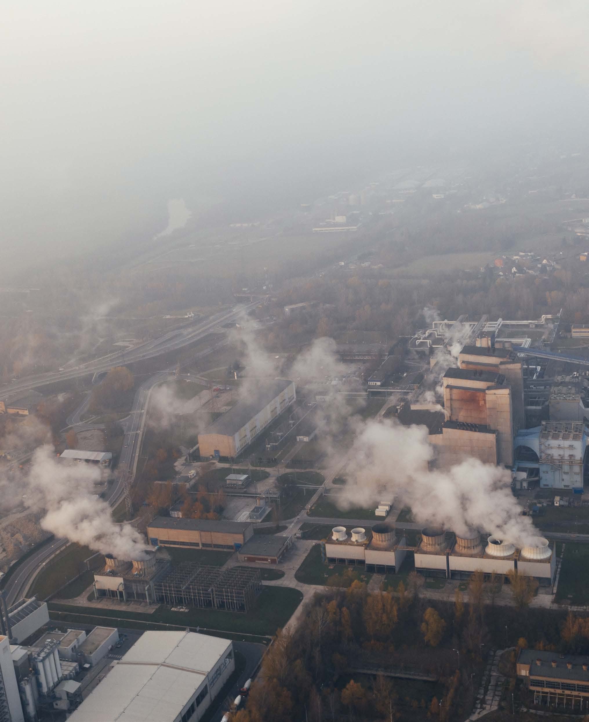 Aerial view of an industrial complex with several buildings, cooling towers emitting plumes of smoke, and roads, indicating industrial activity.